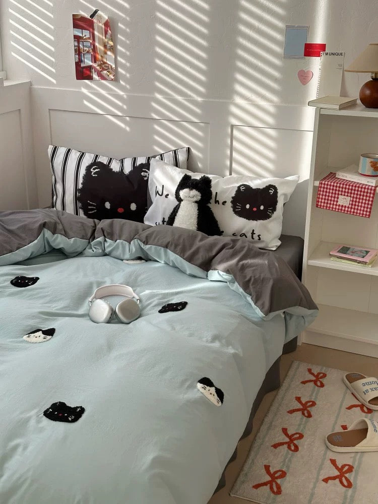 Bed with light blue comforter and cat-themed pillows in a bright room.