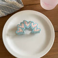 Pink gingham triangle acrylic hair claw clip with tiny blue bow detail, set beside pearls and accessories