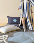 Person wearing a long coat standing next to a bed with pillows and a blue bird design.