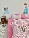 Pink bed with striped bedding in a room setting with a tote bag and small table.