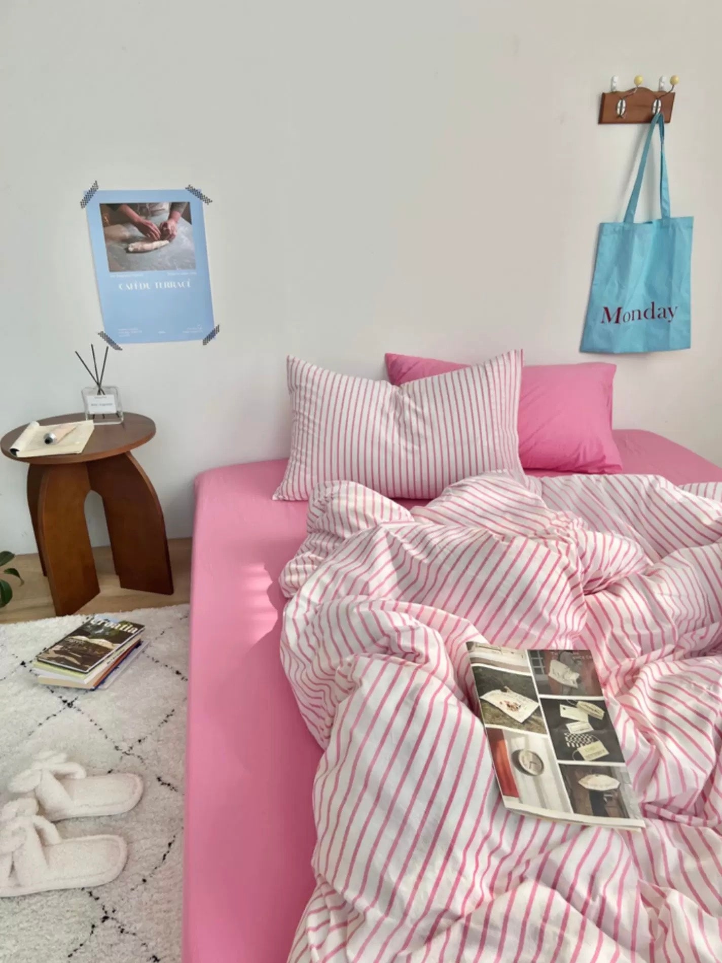 Pink bed with striped bedding in a room setting with a tote bag and small table.