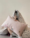 Person sitting on a bed with a pink pillow and a patterned blanket.