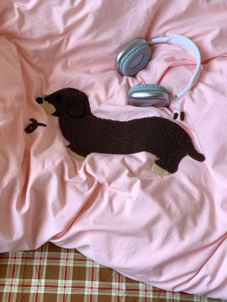 Plush dachshund toy and pink headphones on a pink blanket with plaid fabric underneath.