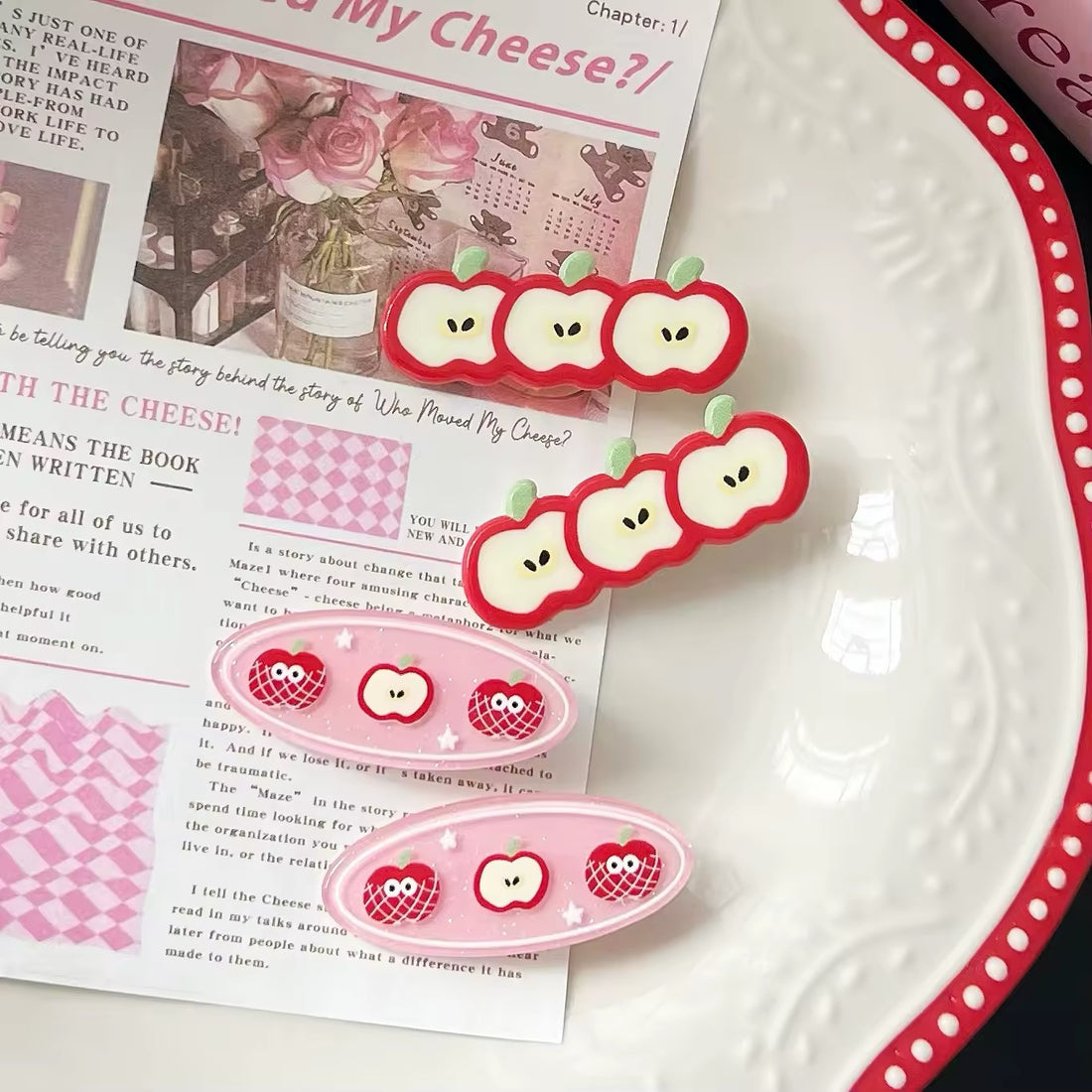 Set of cute apple acrylic hair clips in red and pink tones displayed on a white plate with Korean-style magazine