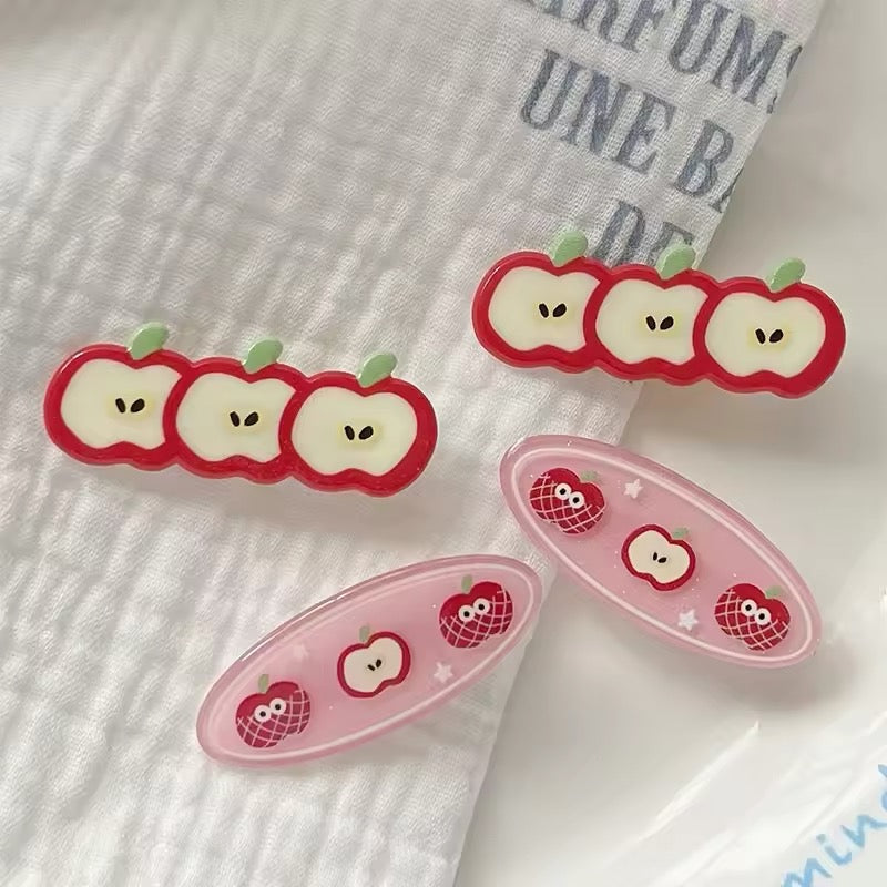 Set of cute apple acrylic hair clips in red and pink tones displayed on a white plate with Korean-style magazine