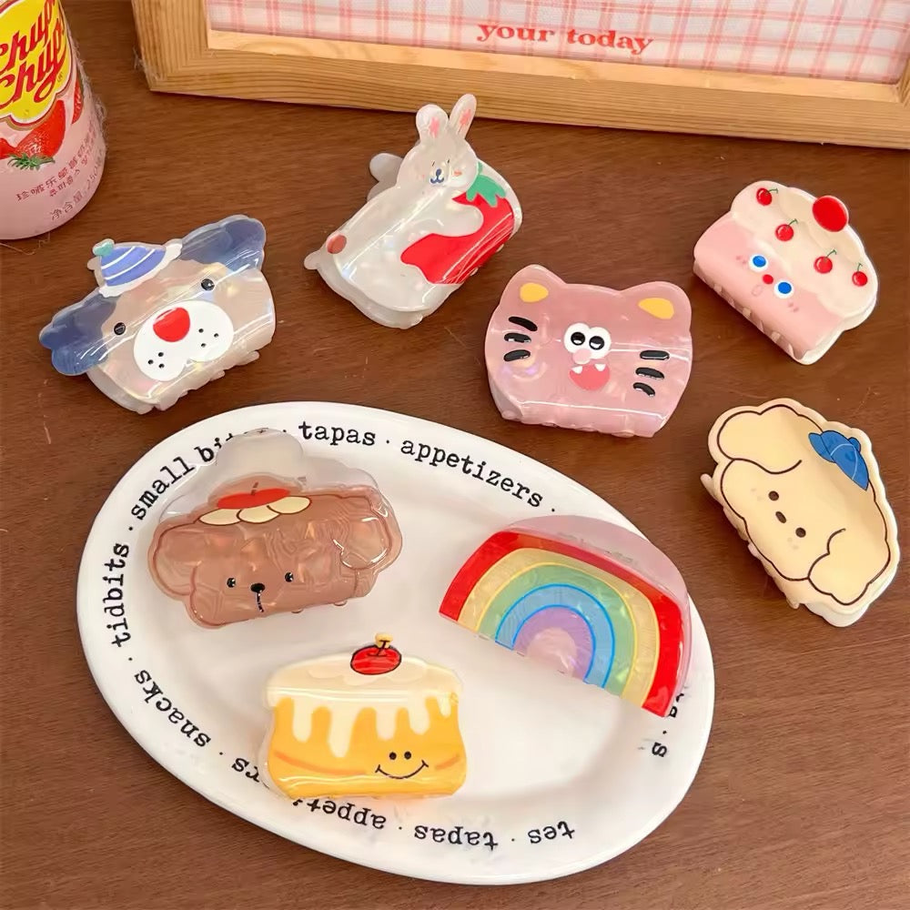 Flat lay of cute cartoon acrylic hair claw clips including rainbow, puppy, cat, and cake designs displayed on a pastel plate