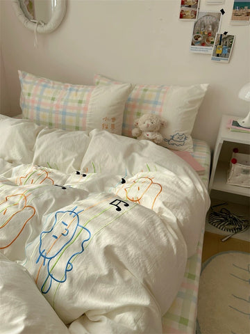 Bed with white comforter featuring colorful cat designs, surrounded by pillows and a teddy bear.