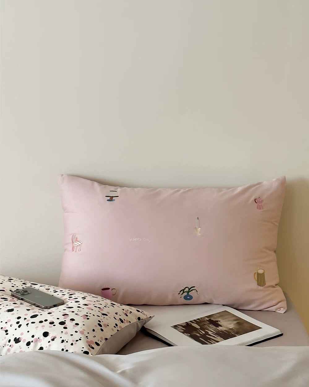 Pink pillow with embroidered designs on a bed with a polka dot blanket and photo album.