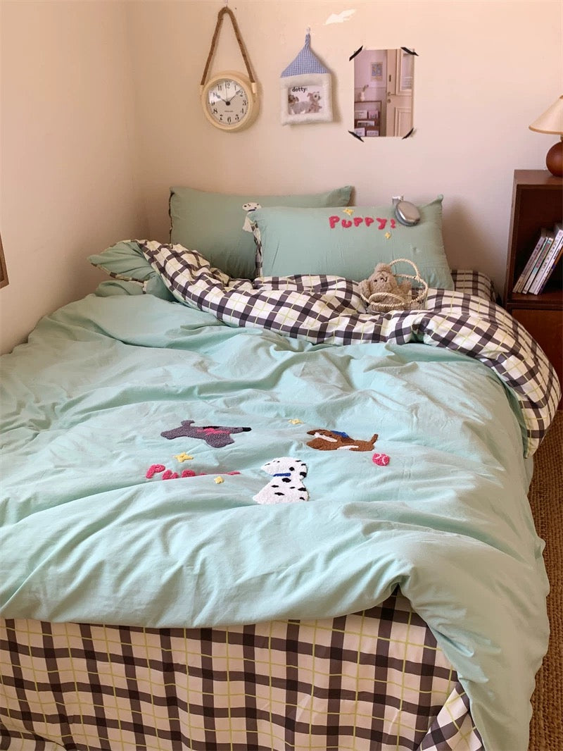 Bedroom with light blue bedding featuring embroidered animals, checkered blanket, and decorative pillows.