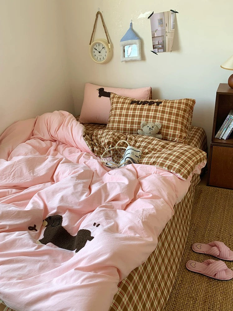 Bed with pink comforter and plaid blanket, pillows, and a teddy bear in a room.