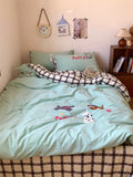 Bed with light blue bedding and checkered blanket, surrounded by decorative items on a shelf.