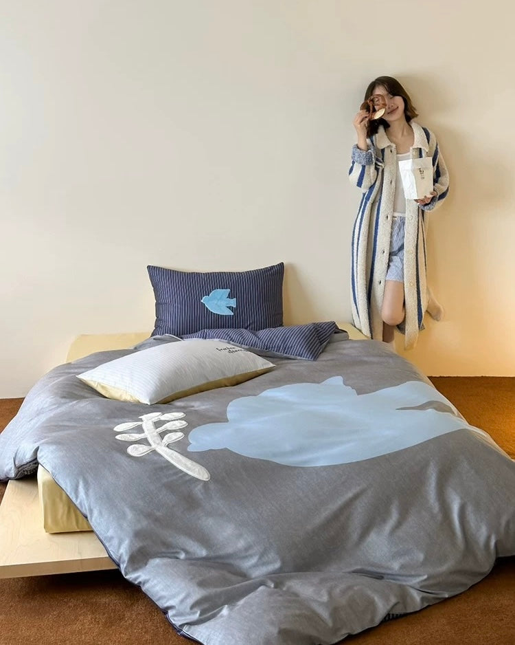 Person standing next to a bed with gray bedding featuring a blue fish design.
