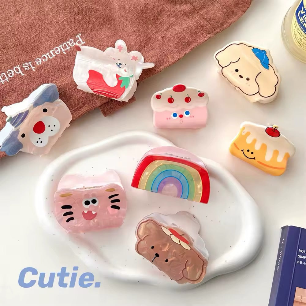 Flat lay of cute cartoon acrylic hair claw clips including rainbow, puppy, cat, and cake designs displayed on a pastel plate