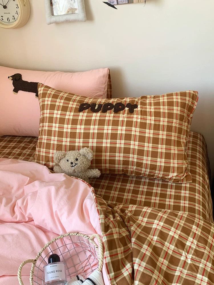 Bed with pink comforter and plaid pillows in a room with a clock and books on a shelf.