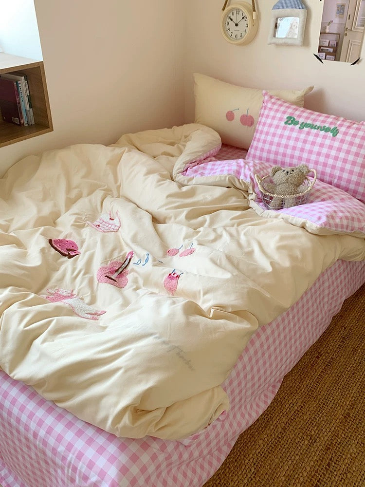Bed with pink checkered sheets and white comforter with floral patterns, in a room setting.
