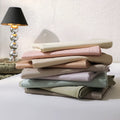 Stack of folded bed sheets in pastel colors on a white surface with a lamp in the background.