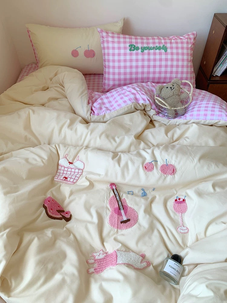 Bed with pink and white checkered pillow, embroidered design, and small teddy bear.