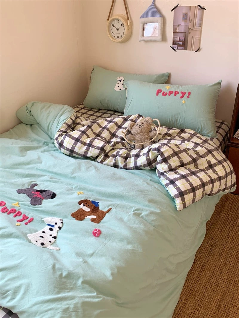 Children's bed with light blue bedding featuring dog appliqués, checkered blanket, and teddy bear.