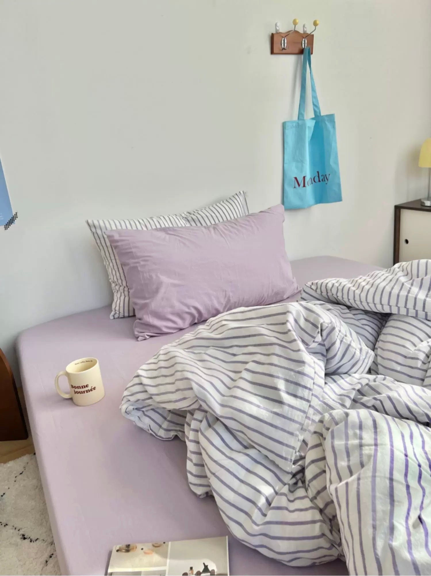 Bed with striped bedding and lavender pillows in a room with a blue bag hanging on the wall.