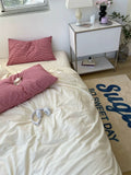 Bedroom with pink pillows on a bed, nightstand with lamp, and 'So Sweet Day' rug.