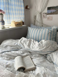 Bedroom with a bed featuring striped pillows and an open book, next to a window with blue plaid curtains.