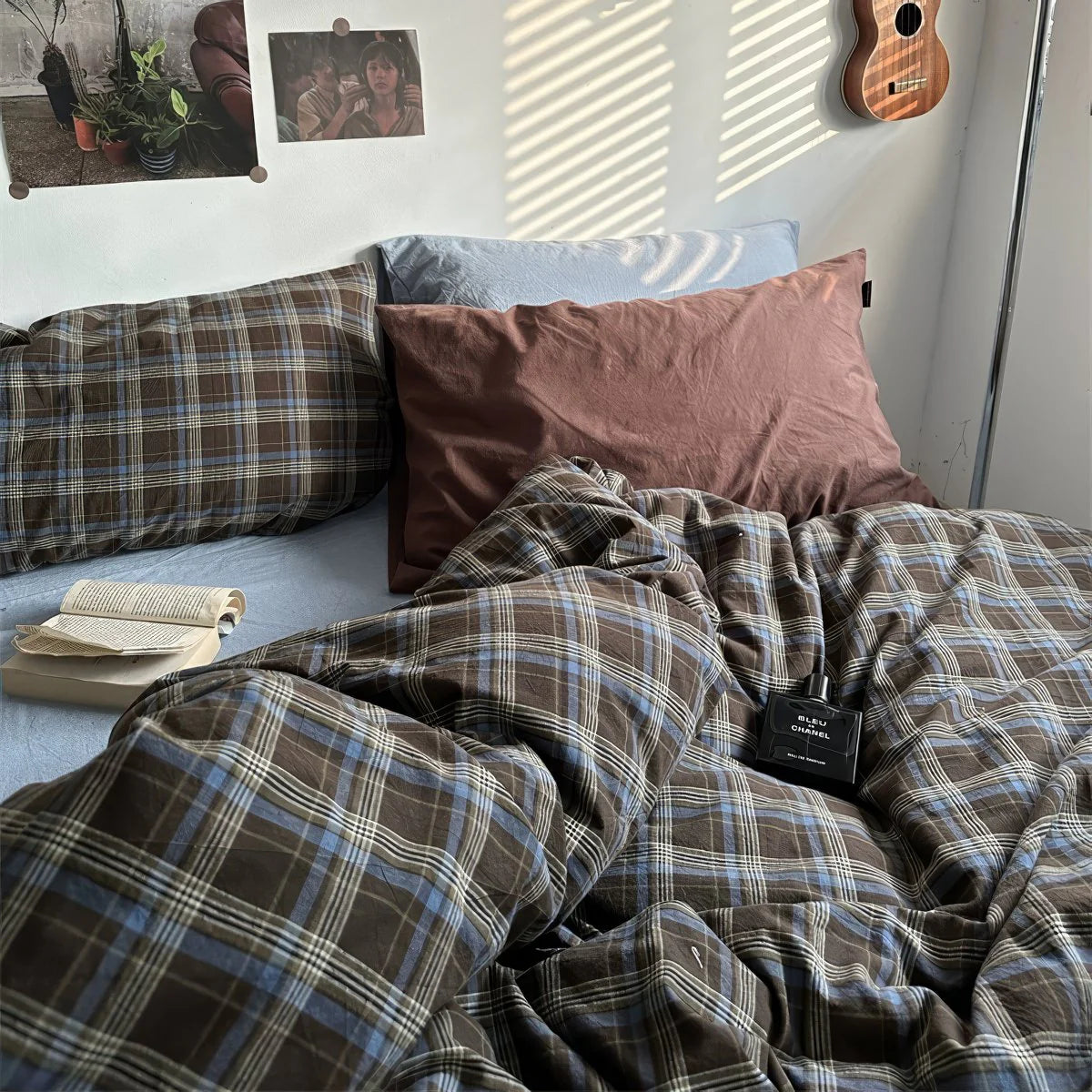 Bed with plaid bedding and pillows, book, and plant in a room.