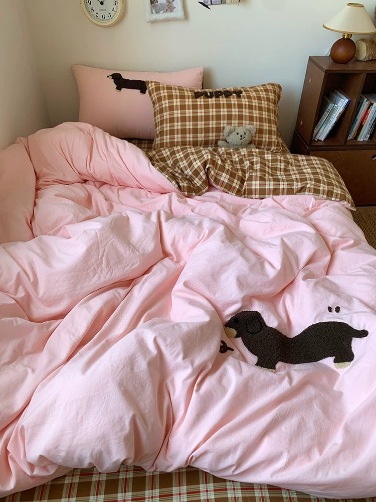 Bed with pink comforter featuring black dog designs, plaid pillows, and a nightstand with books.